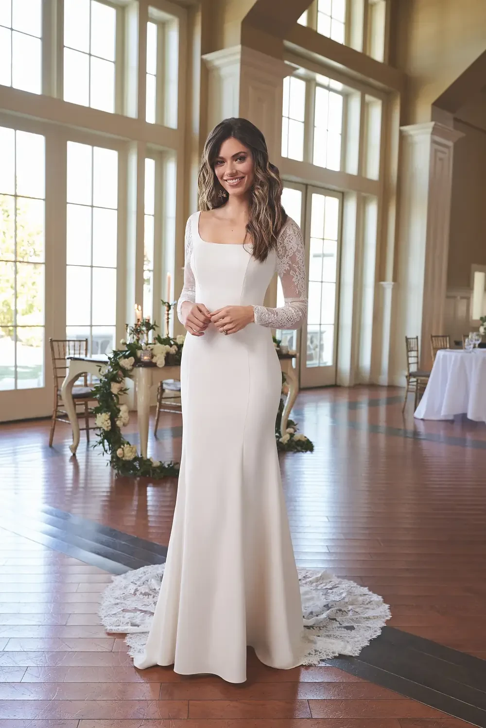 A woman wearing a long-sleeved, form-fitting wedding dress stands in a bright room with large windows, smiling gracefully.