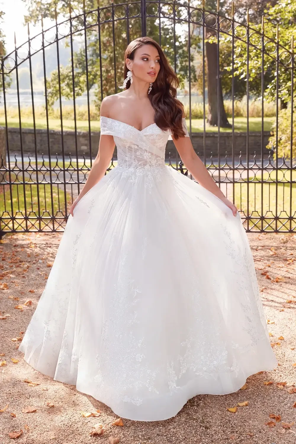 A woman in an elegant white off-the-shoulder gown standing in front of a decorative gate, surrounded by autumn leaves.