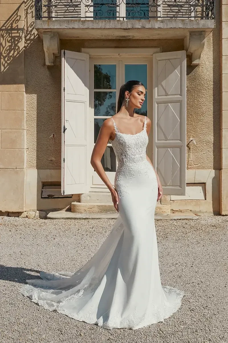 A woman in a fitted white wedding dress with lace details, standing outside a building with large open doors.