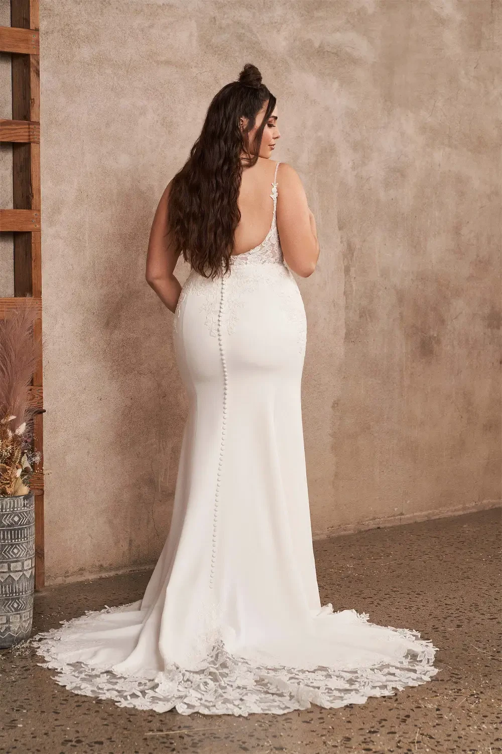A model wearing an elegant, fitted wedding dress with intricate lace detailing, standing confidently in a softly lit, neutral-toned interior.