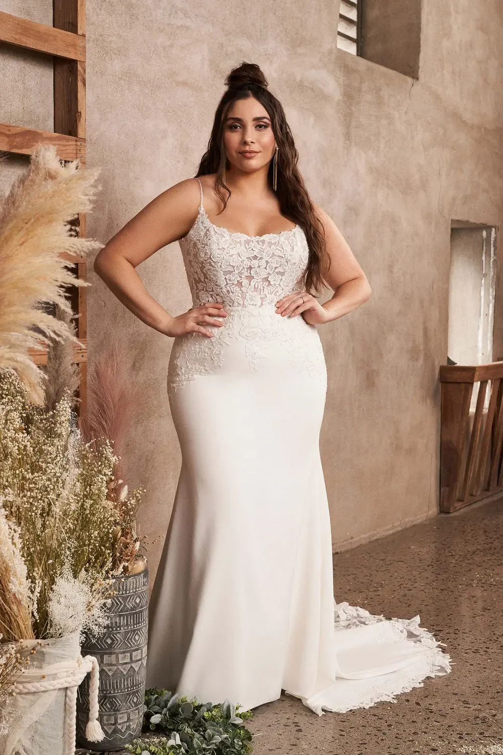 A model wearing an elegant, fitted wedding dress with intricate lace detailing, standing confidently in a softly lit, neutral-toned interior.