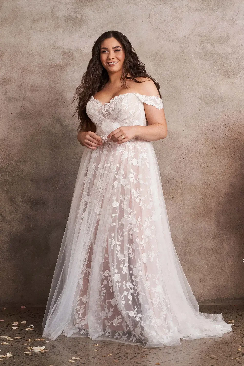 A woman wearing an off-the-shoulder, floral wedding dress standing in front of a textured wall.