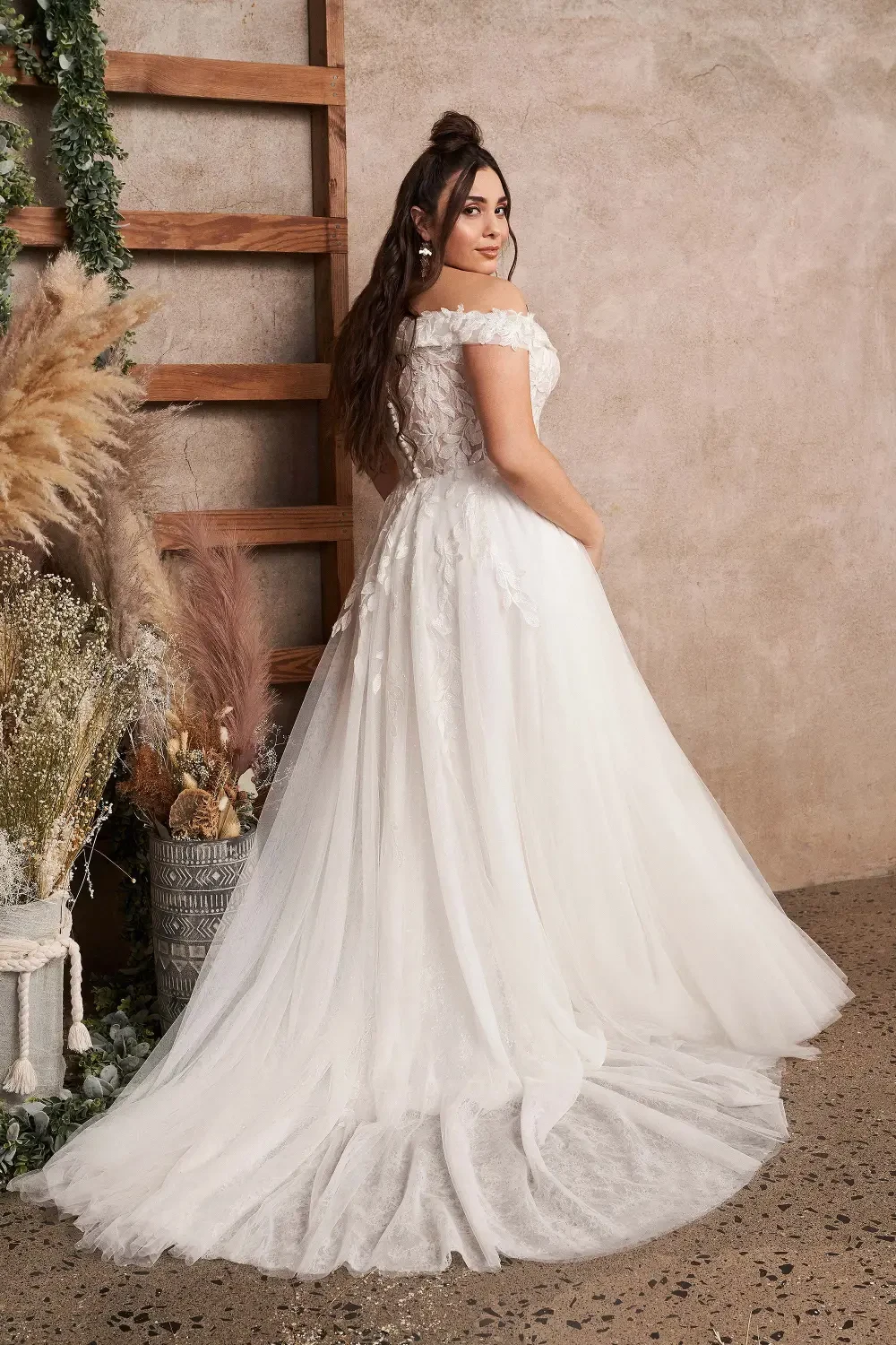 A woman wearing an off-the-shoulder wedding dress, standing with her back to the camera in a softly decorated setting.