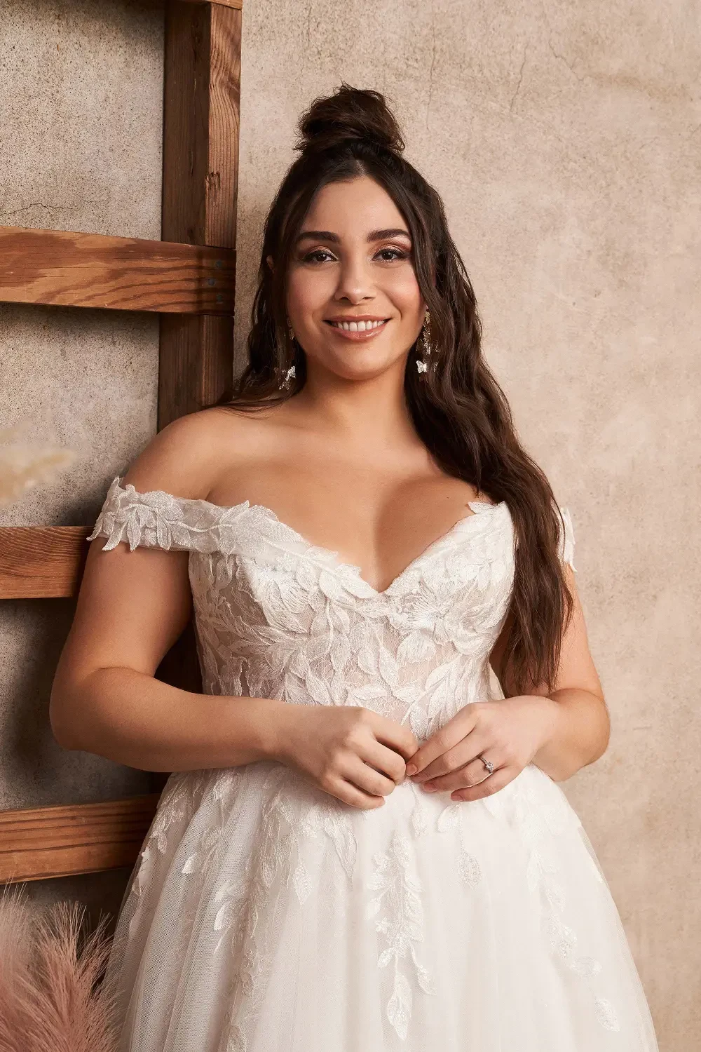 A woman in an elegant off-the-shoulder wedding dress, smiling and posing against a textured background.