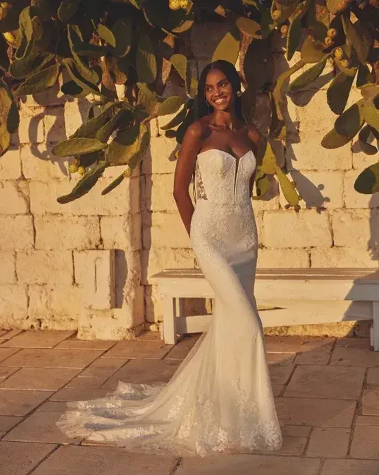 Bride in a strapless, fitted white gown stands joyfully by a sunlit stone wall with leafy cactus backdrop, casting soft shadows.