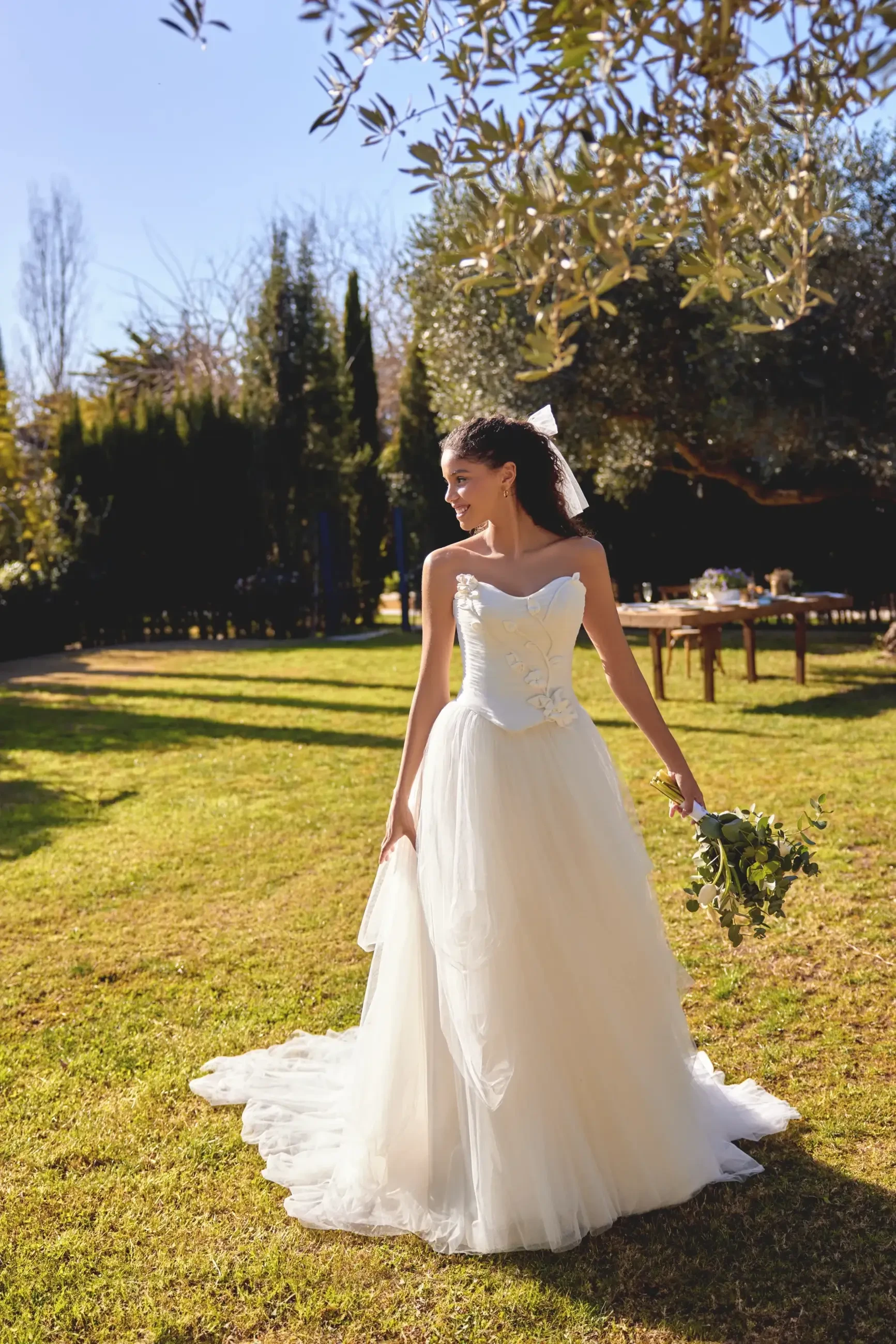 A woman in a white wedding dress holding a bouquet stands in a grassy area with trees in the background.