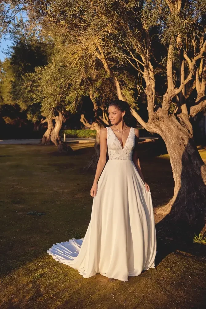 A bride in a flowing white gown stands serenely in a sunlit grove, surrounded by olive trees. The scene conveys tranquility and romance.