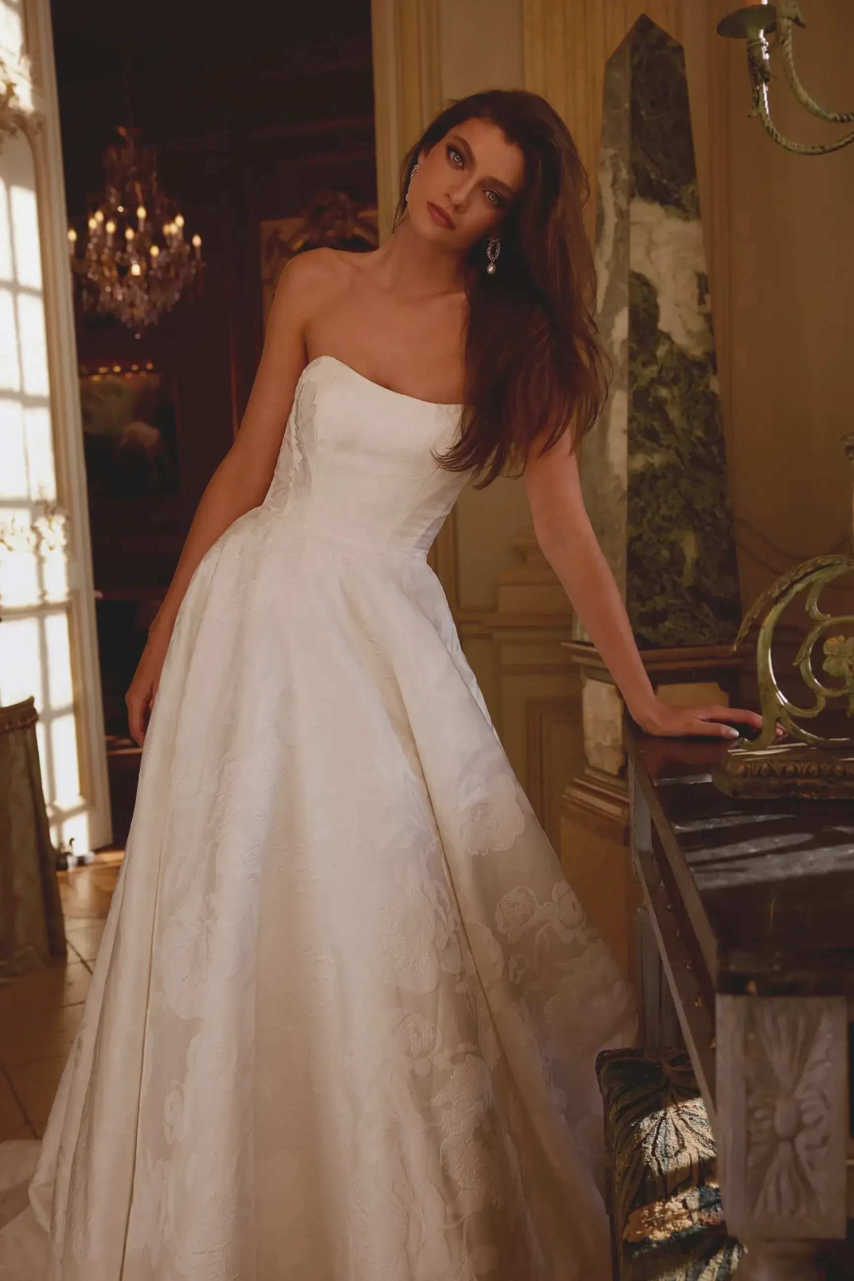 Model is wearing a modern brocade gown featuring floral patterns, chic seaming, a flattering silhouette, and functional pockets.