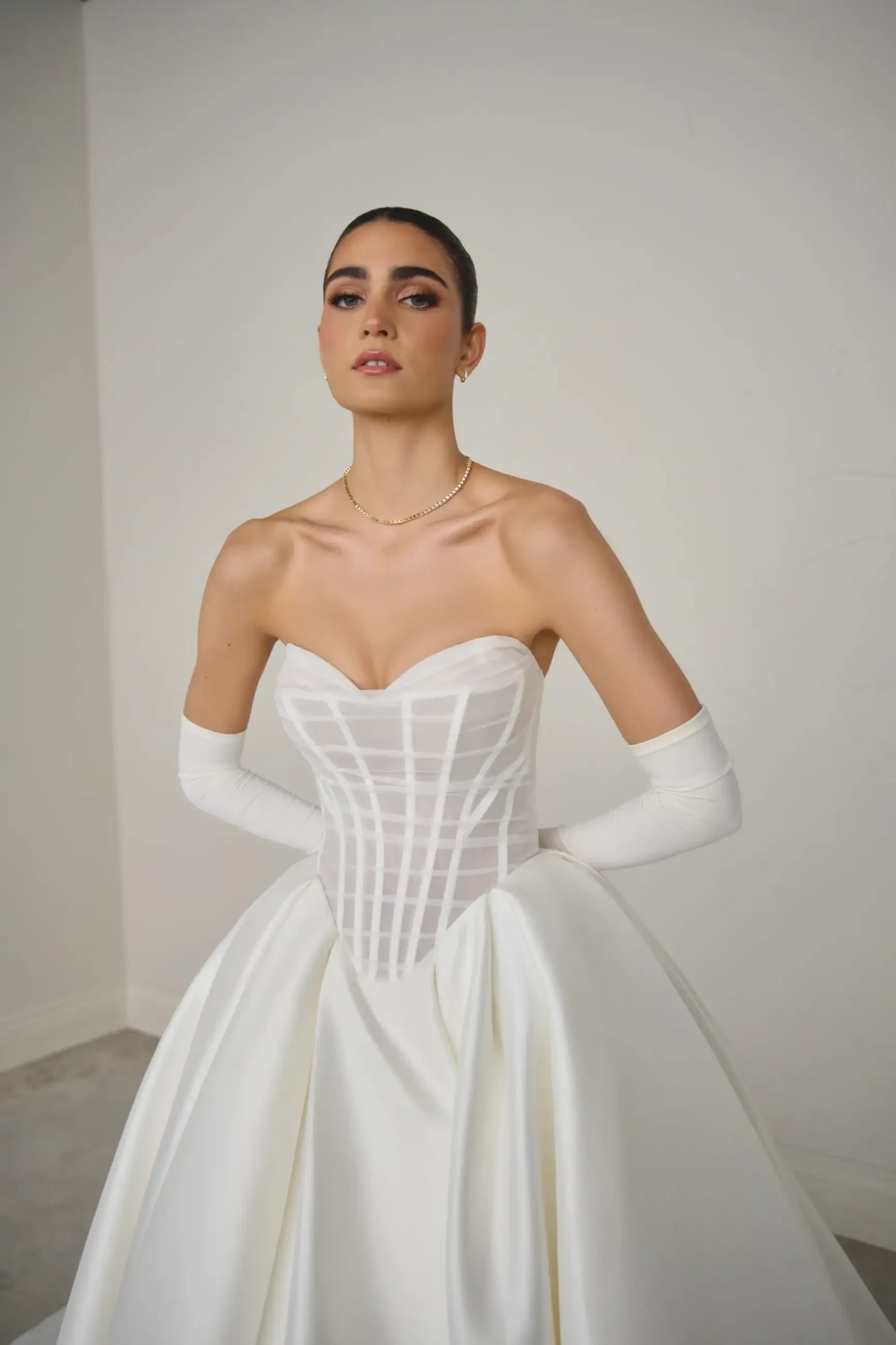 A model wearing a strapless white gown with a fitted bodice and full skirt, styled with long white gloves. The background is a plain light color.