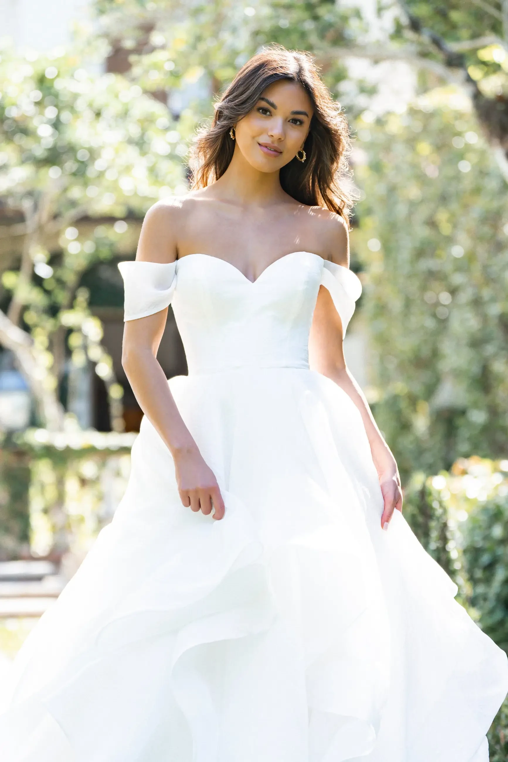 Model is wearing an off-the-shoulder organza gown with a handkerchief hem and layered skirt 