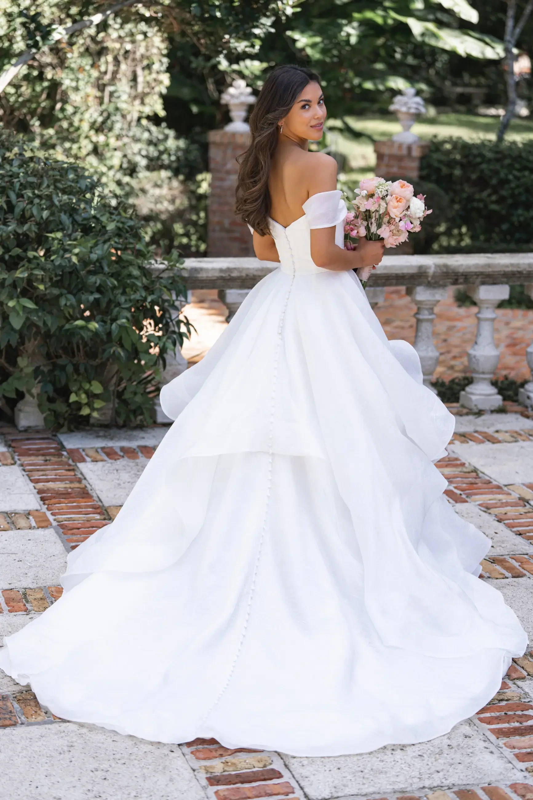 Model is wearing an off-the-shoulder organza gown with a handkerchief hem and layered skirt 