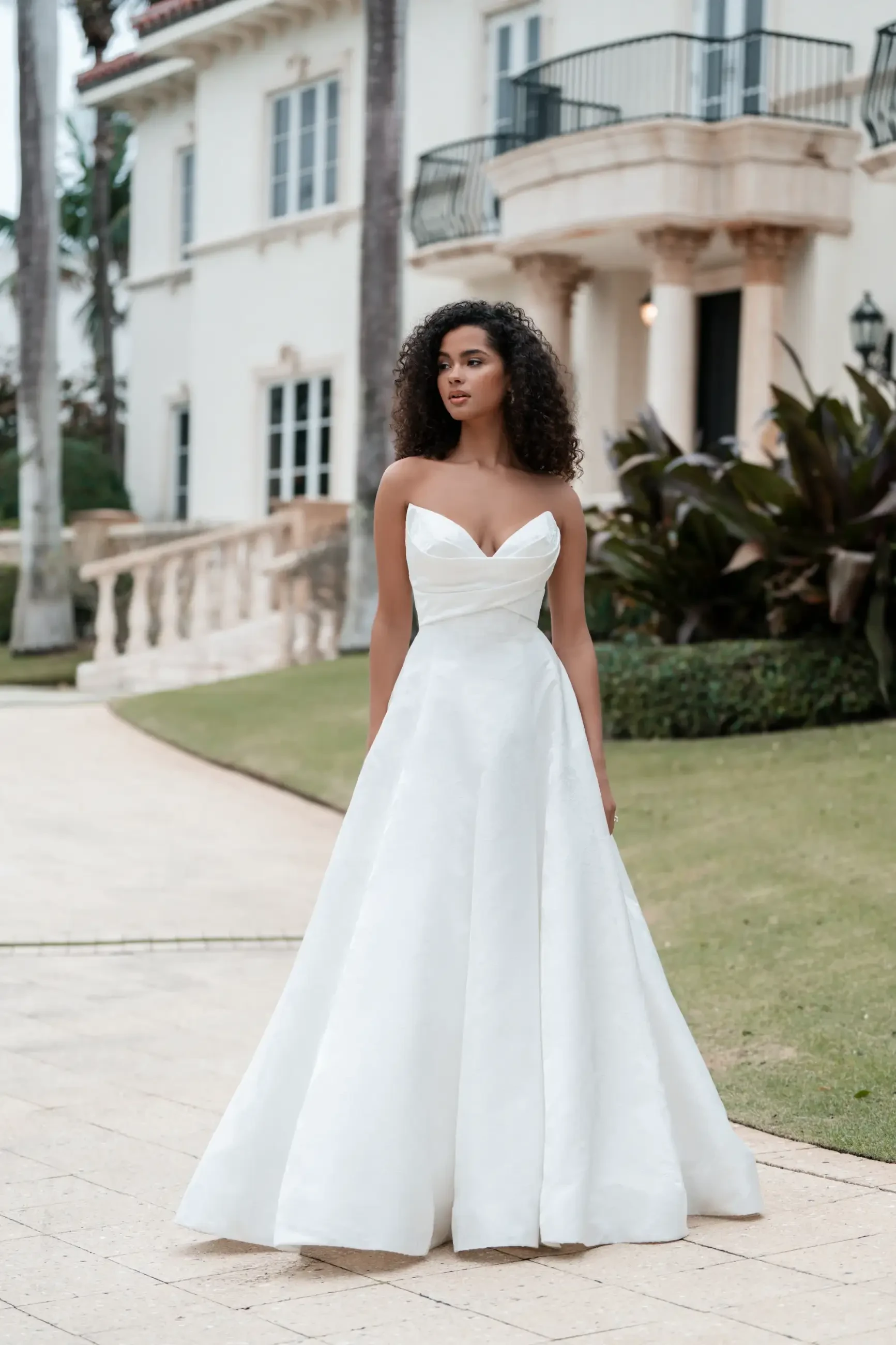 Model is wearing a structured ballgown in opulent brocade with a pointed sweetheart neckline, sculpted bodice, and a dramatic train with covered buttons.