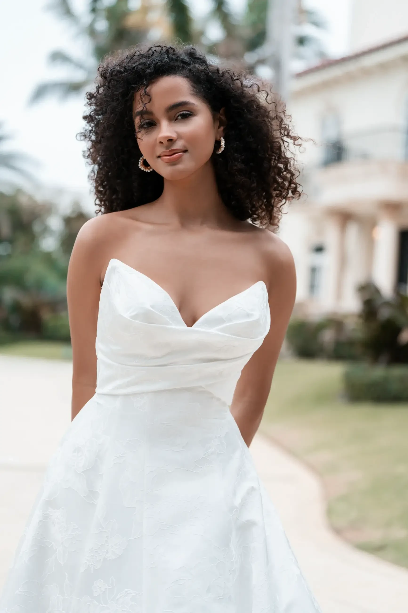 Model is wearing a structured ballgown in opulent brocade with a pointed sweetheart neckline, sculpted bodice, and a dramatic train with covered buttons.