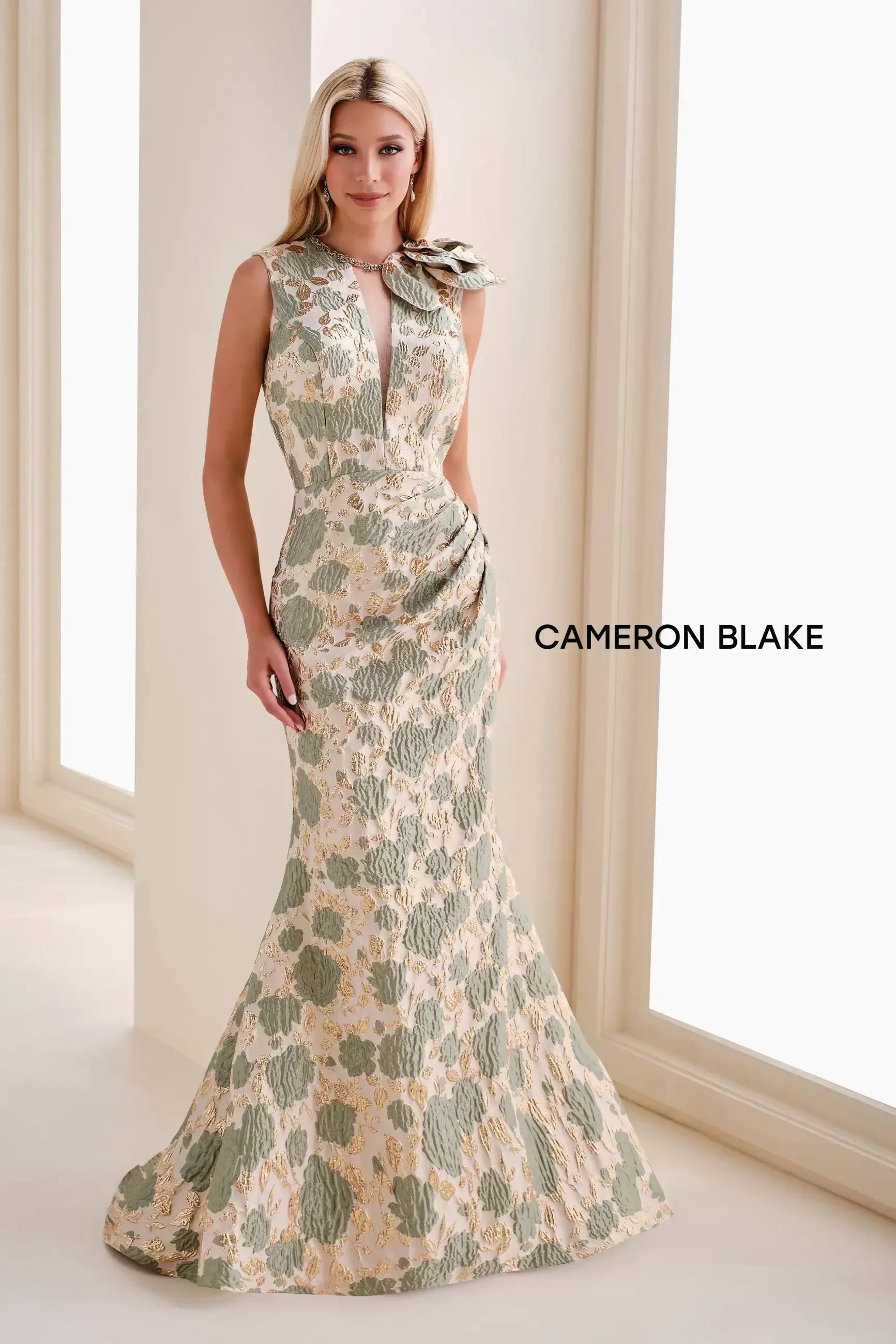 A woman in a sleeveless floral dress with a fitted bodice and flared skirt, standing in a well-lit room. The dress has intricate patterns and includes a bow detail on one shoulder. The name 