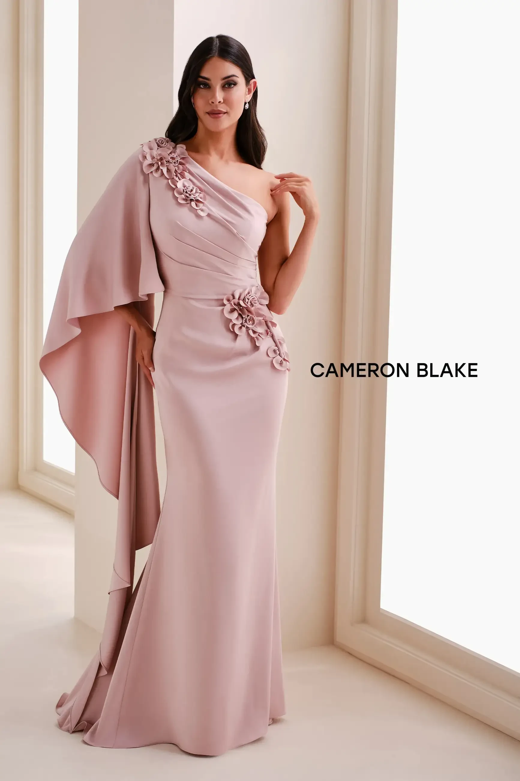 A woman in a blush-colored evening gown with one shoulder draped and floral embellishments, standing by a large window.