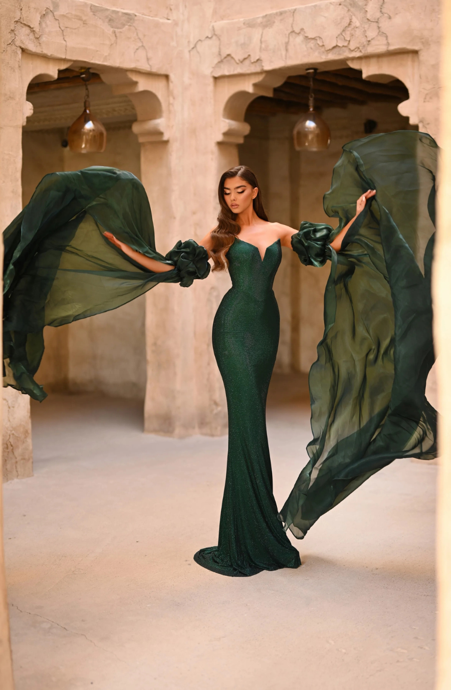 Woman in an elegant, shimmering green gown poses dramatically. Flowing sleeves create a wing-like effect. She stands in an aged, stone-walled room.