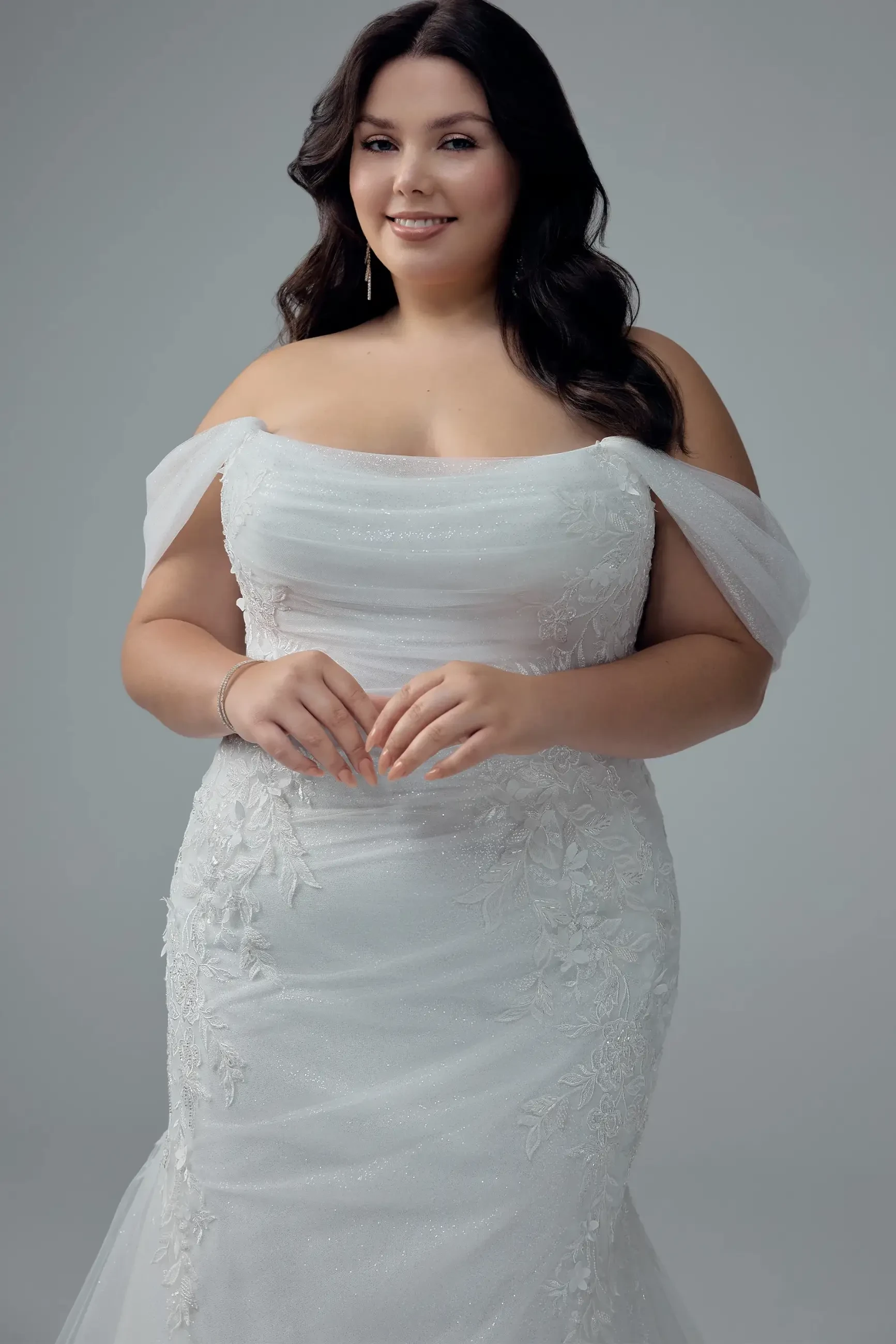 A smiling woman in an off-shoulder, white lace wedding gown stands against a gray background. Her dark hair is styled in soft waves, conveying elegance.